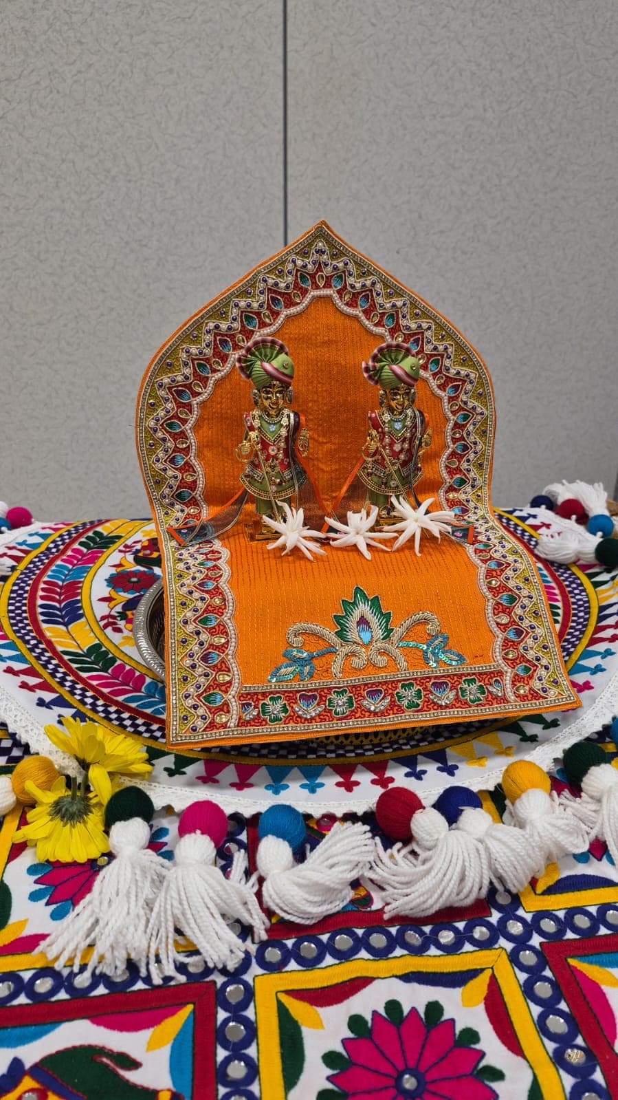 Two murtis on a decorated orange singhasan, with white flowers in front, set on a brightly embroidered cloth.